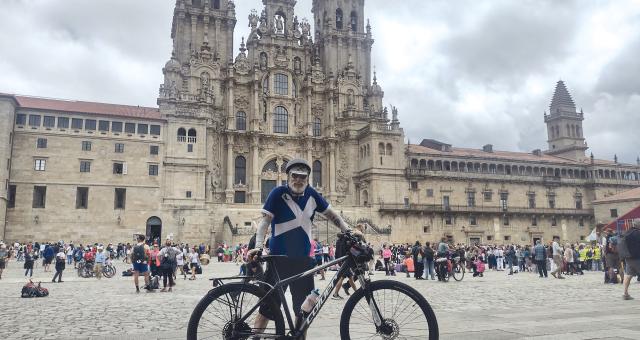 A man in a blue and white cross of St Andrew jersey, black shorts, cycling helmet and sunglasses is standing with a loaded touring bike in front of a cathedral
