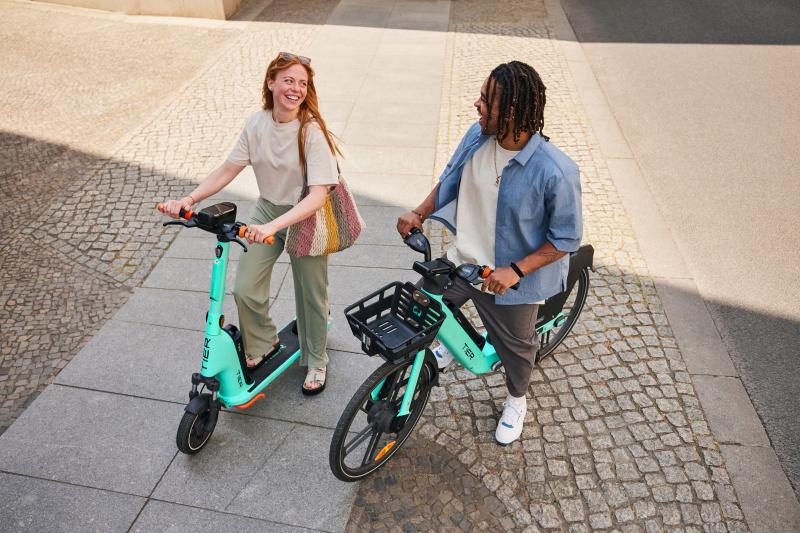 A woman is on a green e-scooter and a man is on a green e-cycle. They are standing still and talking in an urban setting. They are wearing normal clothes. The woman has long red hair, the man had dreadlocks