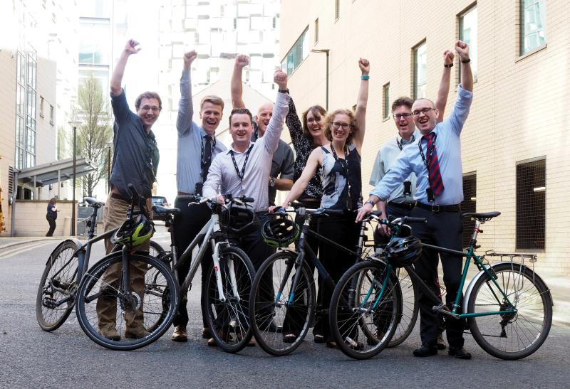 A group of people is standing outside an office building. They are holding bikes and are wearing smart office clothing. They all have one arm in the air as if to celebrate something
