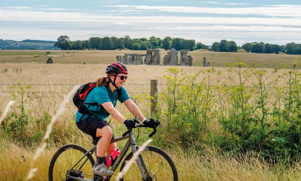 A female cyclists rides past a field in summer with Stonehenge in the background 