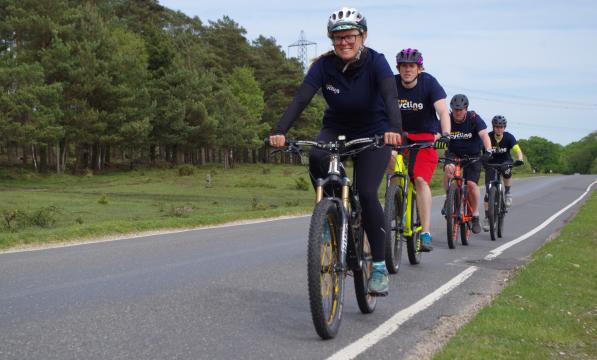Four people are cycling along a quiet country road. All are wearing Cycling UK-branded T-shirts